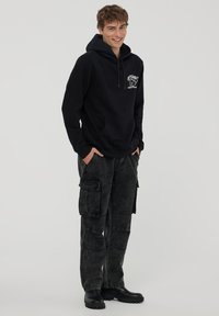 Czarna bluza z kapturem z kieszenią z przodu i graficznym logo; w połączeniu z ciemnoszarymi spodniami cargo z wieloma kieszeniami i luźnym krojem. Czarne buty.