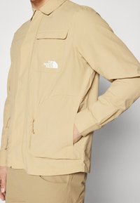 Jasnobeżowa kurtka z lekkiego materiału, z frontowym zamkiem błyskawicznym, dwoma bocznymi kieszeniami oraz kieszenią na piersi z widocznym logo.