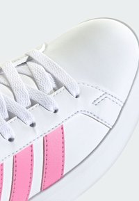 Valkoinen lenkkari valkoisilla nauhoilla ja kahdella pystysuoralla vaaleanpunaisella raidalla sivussa, esitetty lähikuvassa kärkiosasta ja nauhoitusalueelta.