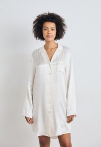 Camicia da notte in satin a maniche lunghe color avorio, con taschino, chiusura a bottoni e motivo testurizzato su tutta la superficie. Tessuto morbido, vestibilità comoda.