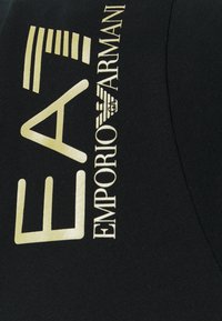 Svart tyg med guldtext och grafiska element. Texten inkluderar "EA7" och "EMPORIO ARMANI" i fet, modern typografi.