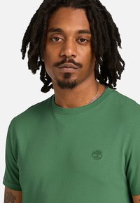 Άνδρας με κοντά dreadlocks και ξυρισμένο μούσι, που φοράει πράσινο μπλουζάκι Timberland με ένα μικρό λογότυπο δέντρου στο στήθος.