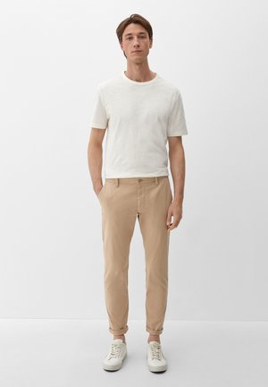 Hellbeige, schmal geschnittene Hose mit glatter Textur und klassischem Knopfverschluss, kombiniert mit einem kurzärmligen, cremefarbenen T-Shirt und weißen Sneakers.