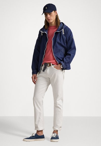 Outfit casual format dintr-o geacă de vânt bleumarin, un tricou roz, pantaloni albi și adidași bleumarin. Detalii notabile includ un centură cu dungi și o șapcă.