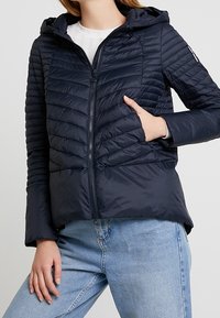 Chaqueta inflable azul marino con capucha, textura acolchada, cremallera frontal y bolsillos laterales, combinada con jeans de color azul claro. Material ligero.