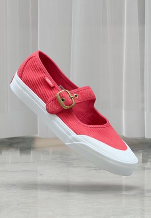 Ballerine Mary Jane en velours côtelé rouge avec boucle dorée, semelle en caoutchouc blanche et étiquette Vans, flottant au-dessus d'un sol réfléchissant devant des stores verticaux.