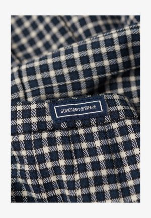 Marineblauw en witte geruite stof met een gestructureerd oppervlak en een zichtbaar "SUPERDAY" merklabel in een rechthoekige blauwe patch.