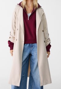 Beige trenchcoat med bred krage och knappstängning, lagd över en burgundy zip-up sweatshirt och ljusblå vida jeans.