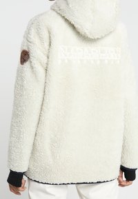 Vit fleece-pullover med huva, med en texturerad yta och broderad logotyp på baksidan, kompletterad med en brun lapp på ärmen.