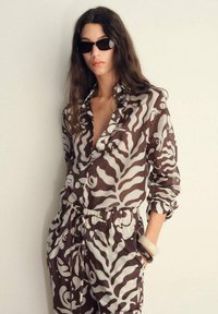 Camicia marrone e crema a fantasia con una vestibilità rilassata e chiusura con bottoni, abbinata a pantaloni coordinati. Il modello indossa occhiali da sole neri.