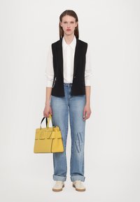 Svart ermeløs vest over hvit skjorte, blå jeans med logo, beige sneakers og en gul håndveske med strukturert design og svarte detaljer.