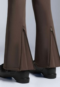 Hnědé rozšířené kalhoty se bočními zipy, vyrobené z hladké, elastické látky. V páru s černou texturovanou obuví, která má moderní design.