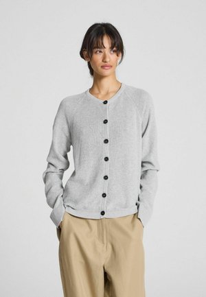 Grijze gebreide cardigan met een ronde halslijn, zwarte knopen en een ribgebreid boord bij de mouwen en onderkant. Gepaard met beige broeken.