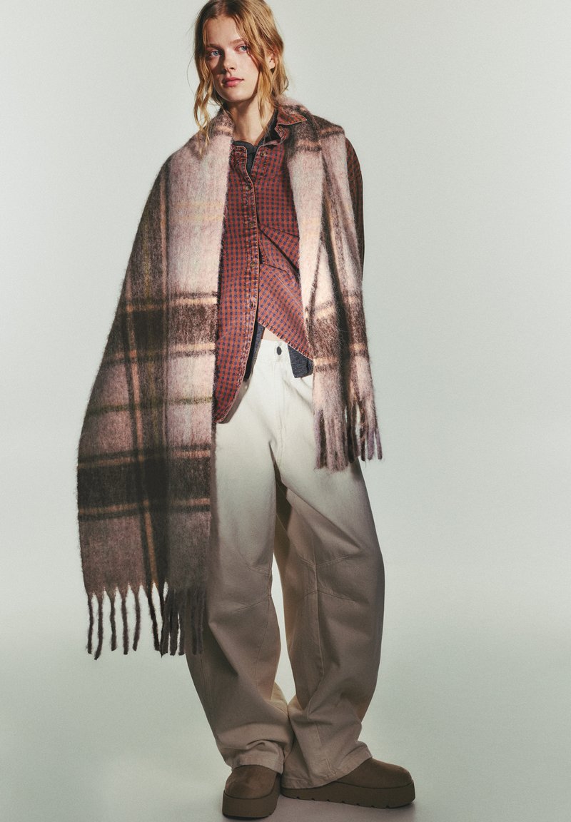 Écharpe en laine à carreaux dans des tons de rose doux, marron et beige, avec des bords à franges. Portée sur une chemise à carreaux rouges et un pantalon blanc ample.