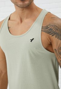 Light green tank top made of soft fabric, featuring a rounded neckline and an embroidered black bird logo on the left chest.