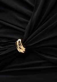 Μαύρο ύφασμα που συγκεντρώνεται και συγκρατείται στη θέση του από ένα λαμπερό, καμπυλωτό χρυσό κλιπ, δημιουργώντας ακτινωτές πτυχώσεις.