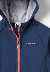 Marineblå jakke med hætte, der har en blød grå fleeceforing og en stærk pink lynlås. Logo vist på venstre frontside.