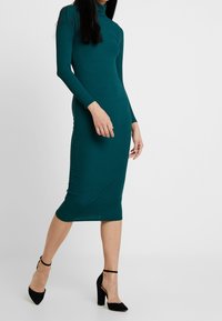 Turkoosin värinen ribattu midi-mekko pitkillä hihoilla ja korkealla kauluksella, jossa on vartalonmyötäinen malli. Yhdistetty mustien korkokenkien kanssa. Minimalistinen tyyli.