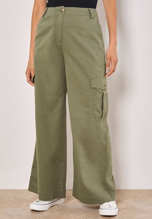 Femme portant un pantalon cargo vert olive à jambes larges avec des poches à boutons, associé à des baskets blanches et un haut noir.