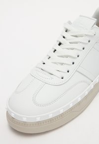 Baskets en cuir blanc avec lacets blancs, couture détaillée, bordure blanche cloutée au-dessus de la semelle beige, vues de trois quarts avant.