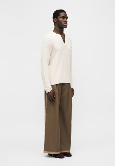 Uomo in piedi, indossa una camicia bianca a maniche lunghe con scollatura a occhiello e pantaloni marroni a gamba larga con strisce laterali beige.