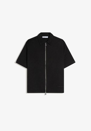 Zwart shirt met korte mouwen, kraag en een zilveren rits die helemaal aan de voorkant loopt, plat gelegd op een witte achtergrond.