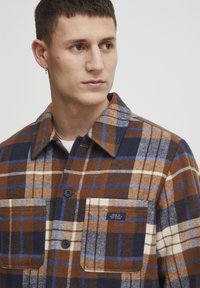 Brown and blue plaid flannel shirt with a collar, two chest pockets, and dark buttons. Fabric has a soft texture and a casual design.