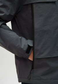 Szara wodoodporna kurtka z elastycznymi mankietami, zapinanymi kieszeniami i gładką fakturą. Posiada zamek błyskawiczny z przodu oraz regulowany linkę napięcia.