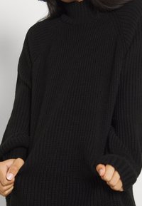 Pull à col roulé en maille noire avec texture côtelée, épaules tombantes et poignets retroussés. Coupe confortable et décontractée, axée sur le confort.