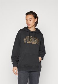 Sweatshirt cu glugă neagră, fabricat din bumbac, cu un buzunar frontal. Designul include cuvântul "TOMMY" în text galben și alb. Croială largă cu manșete ribbate.