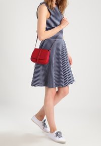 Femme portant une robe sans manches bleu marine et blanche à rayures, chaussée de baskets blanches et portant un petit sac bandoulière rouge, marchant devant un fond uni.