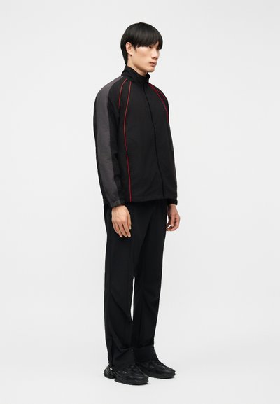 Νεαρός άνδρας όρθιος, φοράει μαύρο αθλητικό μπουφάν με γκρίζα μανίκια και κόκκινο περίγραμμα, μαύρο παντελόνι και μαύρα αθλητικά παπούτσια, σε απλό λευκό φόντο.