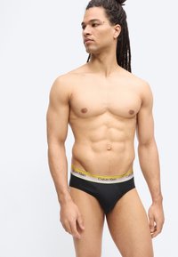 Ευθυτενής άνδρας με μακριά ντραντελός, φορώντας μαύρο εσώρουχο Calvin Klein με κίτρινη μέση, στέκεται μπροστά σε απλό φωτεινό φόντο.