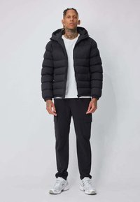 Doudoune noire à capuche, avec des sections matelassées. Portée par-dessus un t-shirt blanc et associée à un pantalon cargo noir et des chaussures grises.