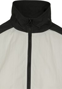 Chaqueta ligera con un cuello negro y acentos en los hombros. Presenta una cremallera frontal completa con tejido texturizado en una combinación de colores blanco y negro.