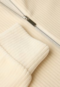 Krämfärgad zip-up-plagg med ribbad textur, slank silverdragkedja och en mjuk, vadderad krage och ärmslut.