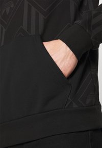 Černá mikina s texturovaným, tónovaným geometrickým vzorem. Má kapsu na břiše a žebrované manžety. Materiál působí měkce a pružně.