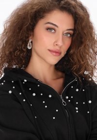 Fekete cipzáras kapucnis pulóver, elöl szétszórt fehér gyöngy díszítéssel. Puha anyag, laza szabás és kapucni.