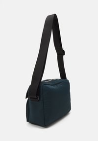Borsa a spalla imbottita di colore teal con tracolla nera regolabile. Presenta una chiusura a zip e una forma rettangolare piatta. Texture liscia e design minimale.
