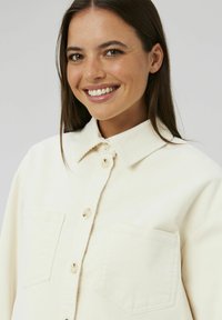 Chemise en velours côtelé crème clair avec un col étalé, fermeture à boutons sur le devant et deux poches poitrine. Présente une texture côtelée et des boutons ronds.