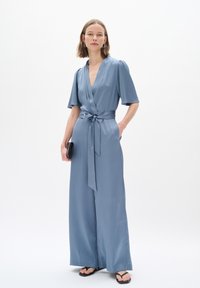 Himmelblaues Wickel-Jumpsuit mit kurzen Ärmeln, Bindegürtel und weitem Bein. Glatte Textur mit subtilen Mustern, accessorisiert mit einer kleinen schwarzen Clutch.