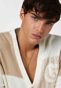 Cardigan en tricot crème et beige avec un col en V, pr�ésentant des panneaux texturés. Le mannequin porte un collier en chaîne doré avec un pendentif.
