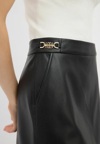 Zwarte leren rok met een slim fit, voorzien van een gouden hardware accent bij de taille en een gladde textuur. Extra stikdetails zichtbaar.