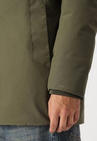 Green, water-resistant jacket with a smooth texture, featuring a side pocket and ribbed cuff. "TECH FUSION" branding visible.