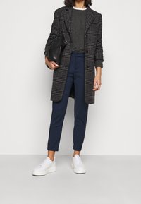 Lungo cappotto a scacchi in nero e grigio, maglione grigio, pantaloni blu navy e sneaker bianche. Tiene una borsa nera. Vestibilità sartoriale.