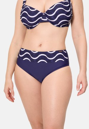 Mujer con un bikini azul marino con rayas onduladas blancas en la parte superior y en la banda de la parte inferior, posando frente a un fondo liso.