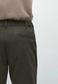 Person trägt dunkelolivfarbene Hosen mit hinterer Paspeltasche und hellbraunen, eingesteckten Pullover, gezeigt von der Taille bis zur Mitte des Oberschenkels vor einfarbigem Hintergrund.