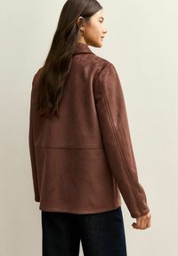 Chaqueta de ante marrón con cuello, mangas largas y textura suave. Presenta un diseño sencillo y costuras a lo largo de la espalda.
