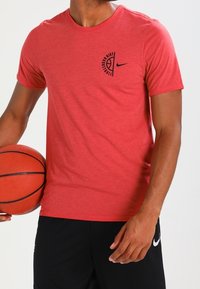 Mężczyzna w czerwonym podkoszulku koszykarskim Nike i czarnych szortach trzyma pomarańczową piłkę do koszykówki na wysokości talii, lekko zwrócony w lewo.