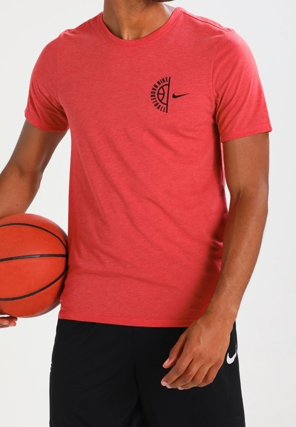 Mężczyzna w czerwonym podkoszulku koszykarskim Nike i czarnych szortach trzyma pomarańczową piłkę do koszykówki na wysokości talii, lekko zwrócony w lewo.